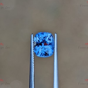 May include: A cushion-cut blue sapphire gemstone held by tweezers. The sapphire is a vibrant blue colour and has a faceted cut.