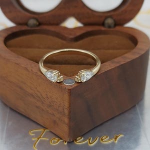 Puede incluir: Un anillo abierto de oro con dos piedras claras en forma de pera en los extremos de la banda. El anillo está colocado en el borde de una caja de madera en forma de corazón con un acabado de veta de madera marrón oscuro y textura lisa.