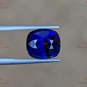 May include: A deep blue cushion-cut gemstone held by tweezers. The gemstone has a faceted surface and is a vibrant blue color.