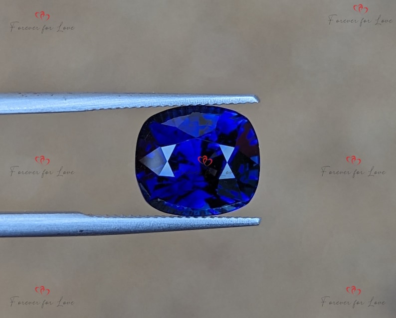May include: A deep blue cushion-cut gemstone held by tweezers. The gemstone has a faceted surface and is a vibrant blue color.