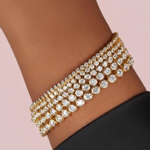 Puede incluir: Un brazalete de oro con m&uacute;ltiples filas de piedras preciosas redondas y transparentes. El brazalete est&aacute; engarzado en una banda de oro y se lleva en la mu&ntilde;eca. El fondo es de color rosa claro.