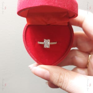 May include: A gold engagement ring with a large rectangular diamond in a red velvet heart-shaped box.