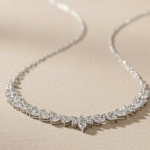 Puede incluir: Un collar plateado con una cadena delicada y una serie de piedras preciosas de talla marquesa que forman un dise&ntilde;o curvo. Las piedras est&aacute;n muy juntas, creando una apariencia brillante y elegante. El collar se muestra sobre una superficie de tono neutro.