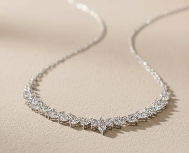 Puede incluir: Un collar plateado con una cadena delicada y una serie de piedras preciosas de talla marquesa que forman un dise&ntilde;o curvo. Las piedras est&aacute;n muy juntas, creando una apariencia brillante y elegante. El collar se muestra sobre una superficie de tono neutro.