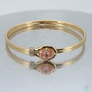 Puede incluir: Una pulsera dorada con una gema ovalada de color melocotón en el centro. La pulsera tiene un cierre de bisagra y está adornada con pequeñas piedras transparentes en un lado del engaste de la gema. La pulsera se muestra sobre una superficie reflectante.