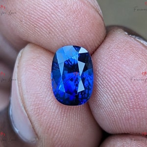 May include: A vibrant blue oval-shaped gemstone with a faceted cut. The gemstone is held between two fingers.