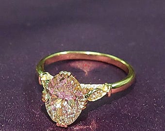 May include: A gold engagement ring with an oval-shaped diamond set in a delicate, ornate setting. The diamond has a slight pink hue and is surrounded by smaller diamonds.