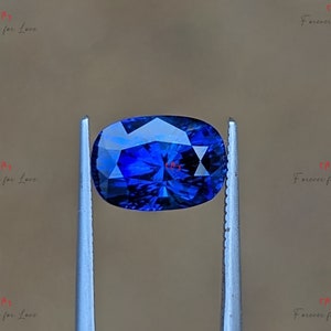 May include: A deep blue oval sapphire gemstone held in tweezers. The sapphire has a faceted cut and a brilliant shine.