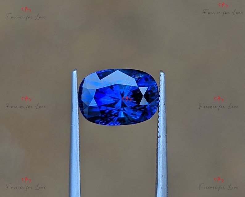 May include: A deep blue oval sapphire gemstone held in tweezers. The sapphire has a faceted cut and a brilliant shine.