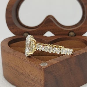 May include: A gold engagement ring with a large, clear, oval-cut center stone and a band lined with smaller, oval-cut stones. The ring is set in a wooden heart-shaped box.