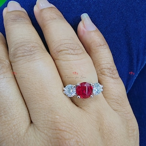 May include: A silver ring with a large red gemstone surrounded by smaller clear gemstones. The ring is on a person's finger.