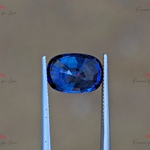 May include: A deep blue oval-shaped gemstone held by tweezers. The gemstone has a faceted cut and is a vibrant blue color.