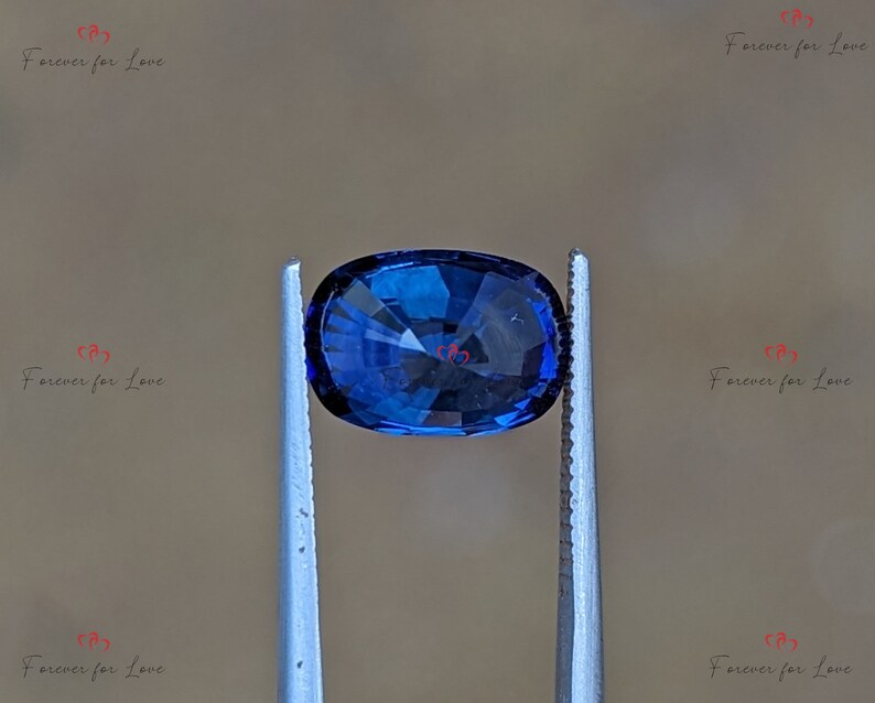 May include: A deep blue oval-shaped gemstone held by tweezers. The gemstone has a faceted cut and is a vibrant blue color.