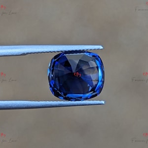 May include: A deep blue gemstone with a cushion cut, held in tweezers against a light brown background.