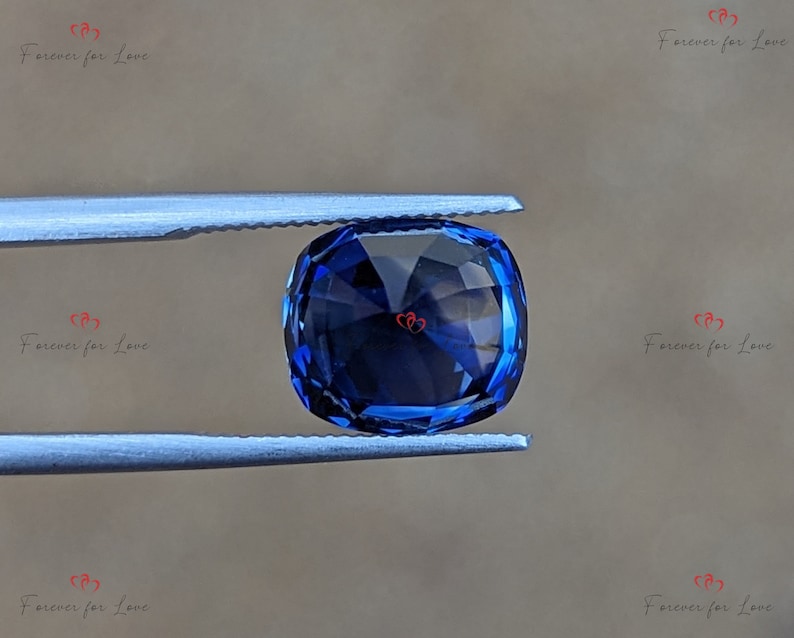 May include: A deep blue gemstone with a cushion cut, held in tweezers against a light brown background.
