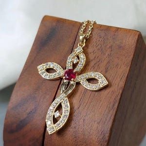 May include: A gold-colored pendant necklace featuring a cross design. The cross is adorned with small, sparkling diamonds and a central, vibrant red gemstone. The necklace is displayed on a wooden surface.