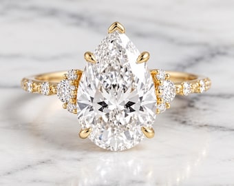 May include: A gold ring featuring a large pear-shaped center stone held by five prongs. The band is crafted from yellow gold and is accented with small round stones on both sides of the center setting, extending along the shank of the ring.