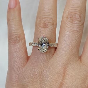 May include: A gold ring featuring an oval-cut diamond center stone. The band is adorned with smaller diamonds. The ring is shown on a person's finger, showcasing the ring's sparkle and design.
