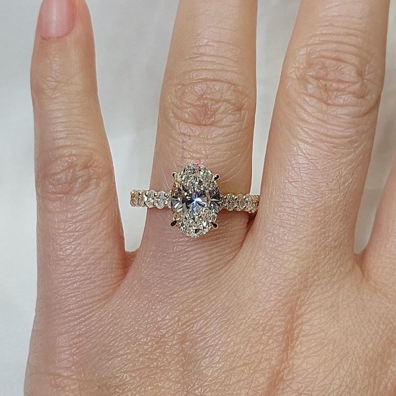 May include: A gold ring featuring an oval-cut diamond center stone. The band is adorned with smaller diamonds. The ring is shown on a person's finger, showcasing the ring's sparkle and design.