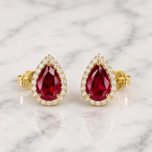 Peut inclure: Une paire de boucles d'oreilles à clous en or avec une pierre précieuse rouge en forme de poire au centre, entourée d'un halo de petites pierres blanches rondes. Les boucles d'oreilles présentent un sertissage à griffes et une tige arrière.