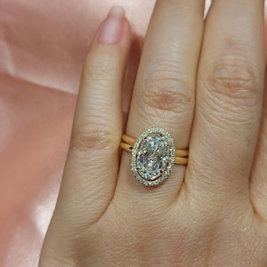 May include: A gold engagement ring with a large oval-shaped diamond surrounded by a halo of smaller diamonds. The ring is on a finger.