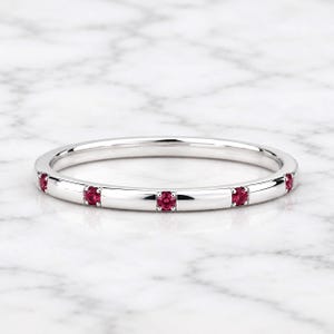 May include: A thin, polished silver-toned metal band ring featuring five small, round, vibrant pinkish-red gemstones set at equal intervals along the outer circumference of the band. The ring has a smooth, reflective finish and a minimalist design.
