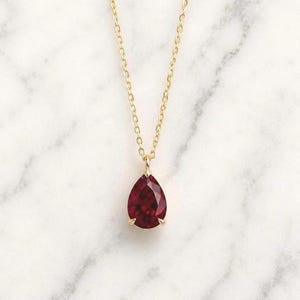 Op de afbeelding: Een verfijnde gouden ketting met een enkele peervormige, dieprode edelsteen hanger. De gefacetteerde steen is bevestigd in een eenvoudige gouden zetting met pootjes en hangt aan een dunne, gepolijste gouden schakelketting die verticaal naar beneden valt.