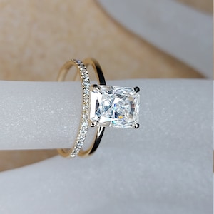 May include: A radiant-cut diamond engagement ring with a gold band, paired with a delicate gold band adorned with small, sparkling stones. The rings are set against a soft, neutral background, highlighting the brilliance of the diamonds.