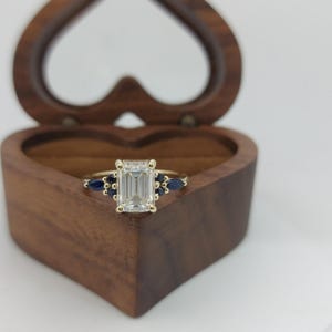 Puede incluir: Un anillo de oro con un diamante de talla esmeralda y acentos de zafiro azul. El anillo está en una caja de madera en forma de corazón.