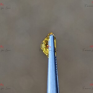 May include: A close-up of a yellow gemstone held by tweezers. The gemstone is faceted and has a golden hue.