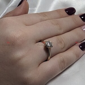 May include: A gold engagement ring with a round, clear diamond center stone. The ring is on a person's finger.