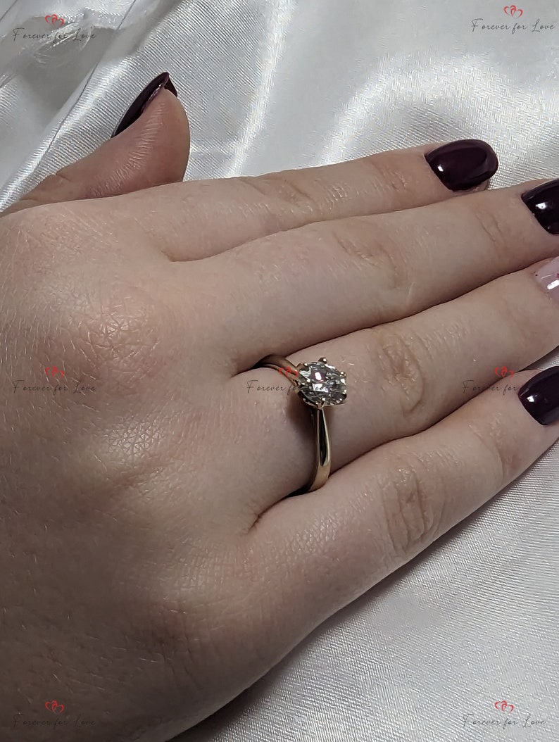 May include: A gold engagement ring with a round, clear diamond center stone. The ring is on a person's finger.