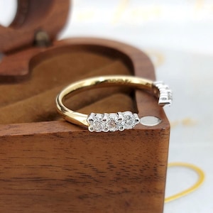 Puede incluir: Un anillo de oro y diamantes con tres diamantes prominentes en fila. El anillo se encuentra sobre un joyero de madera, resaltando su elegante diseño y brillo. La banda del anillo es dorada y los diamantes son claros.