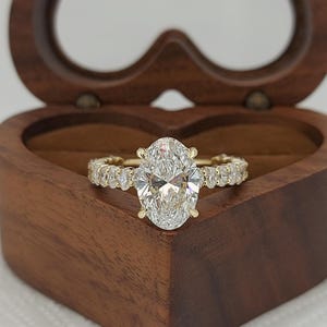 May include: A gold ring with a large oval-cut diamond center stone and smaller diamonds along the band. The ring is displayed inside a heart-shaped wooden box with a dark brown finish. The ring is a piece of jewelry.