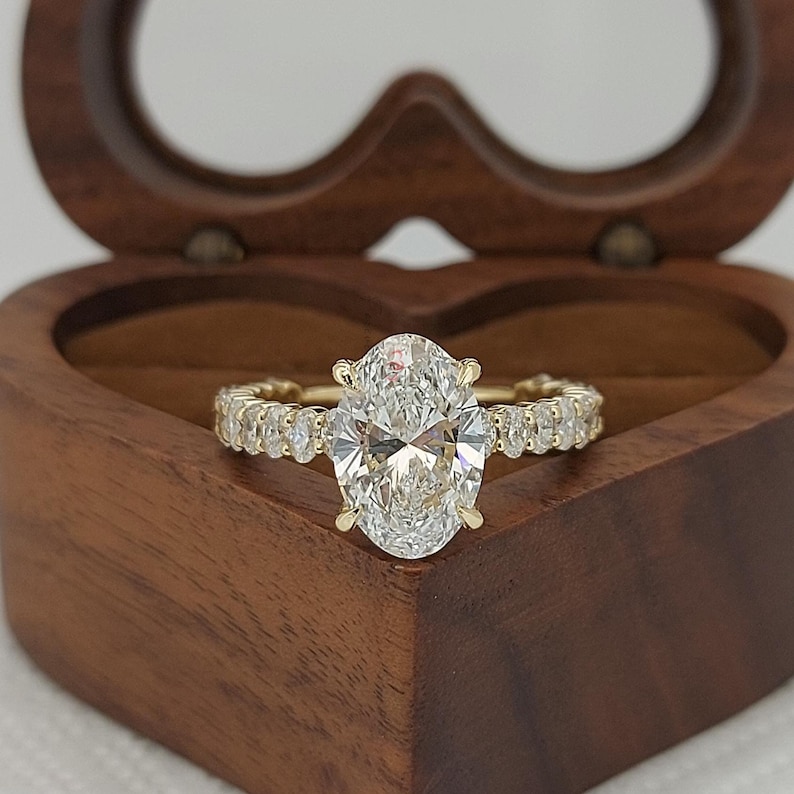 May include: A gold ring with a large oval-cut diamond center stone and smaller diamonds along the band. The ring is displayed inside a heart-shaped wooden box with a dark brown finish. The ring is a piece of jewelry.