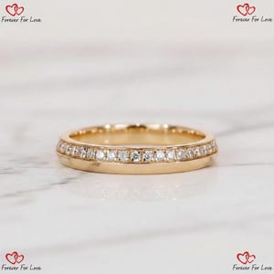 May include: A gold band ring with a double row of small, clear gemstones. The ring is set on a white marble surface. The ring is a wedding band or engagement ring.