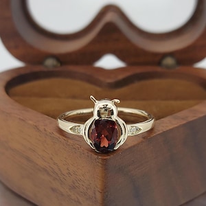 Pode incluir: Um anel dourado com um design de coruja estilizado. O corpo da coruja é uma gema grande, oval, de cor vermelha escura, com pequenos detalhes de diamante em ambos os lados. O anel está em uma caixa de madeira em forma de coração.