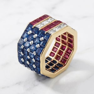 May include: A gold ring with a design reminiscent of the American flag. The ring features blue sapphires, red rubies, and white diamonds. The ring has an octagonal shape and is set on a white surface.
