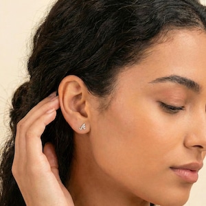 May include: A close-up of a person's ear with a small, silver stud earring in the shape of the letter "A". The earring is set with small, sparkling stones. The person's skin is a light, warm tone, and their dark hair is out of focus in the background.