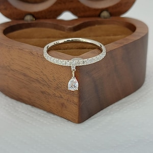 Könnte beinhalten: Ein silberner Ring mit einem Band, das mit kleinen, klaren Steinen besetzt ist. Ein tropfenförmiger klarer Stein hängt von der Mitte des Rings. Der Ring wird in einer herzförmigen Holzbox präsentiert.
