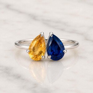 Könnte beinhalten: Ein silberfarbener Ring mit zwei tropfenförmig geschliffenen Edelsteinen, die nebeneinander angeordnet sind. Ein Stein ist leuchtend gelb, der andere tiefblau. Die Steine werden von kleinen Metallfassungen auf einer glatten Ringschiene gehalten.