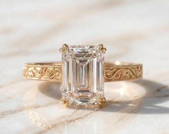 Op de afbeelding: Een gouden verlovingsring met een smaragdgeslepen diamant. De ring heeft een gegraveerde band en een vier-klauwzetting. De diamant is helder en reflecteert licht, op een marmeren oppervlak.