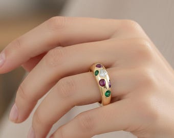 Può includere: Un anello a fascia in oro con una serie di pietre preziose incastonate a filo, tra cui pietre rotonde viola, verdi e una singola pietra trasparente a forma di aquilone. La superficie in oro lucido ha un profilo liscio e arrotondato.