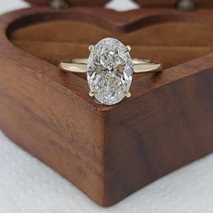 May include: A gold engagement ring with an oval-cut diamond. The ring is set in a four-prong setting and is displayed in a wooden heart-shaped box. The diamond sparkles, reflecting light.