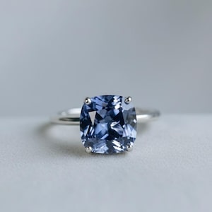 May include: A silver ring with a large, square-cut, light blue gemstone. The gemstone is secured by four prongs. The ring is set against a soft grey background, emphasising the ring's elegant design.