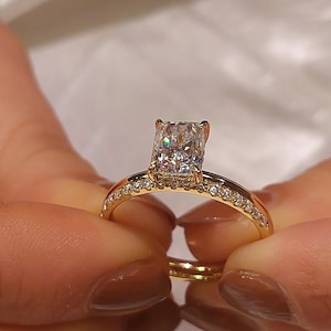 May include: A gold engagement ring with a large, square-shaped diamond. The ring has a band of small diamonds.