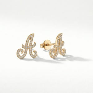 May include: A pair of gold-colored stud earrings shaped like the letter "A". The earrings are encrusted with small, clear gemstones. The earrings are displayed on a white surface.