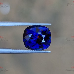 May include: A deep blue cushion-cut gemstone held in tweezers. The gemstone has a faceted surface and is a vibrant blue color.