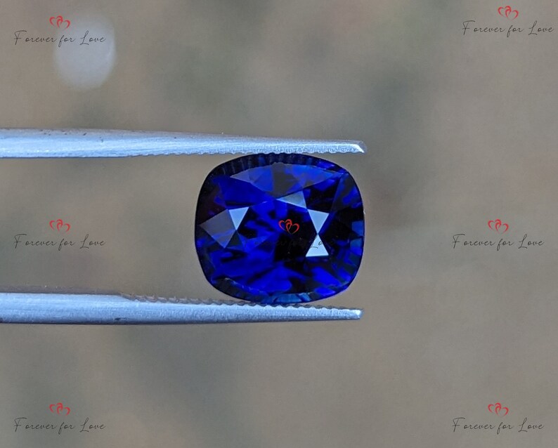 May include: A deep blue cushion-cut gemstone held in tweezers. The gemstone has a faceted surface and is a vibrant blue color.