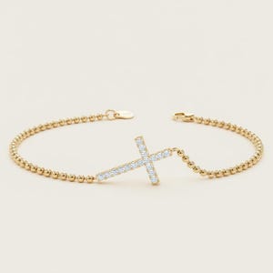 May include: A gold beaded chain bracelet featuring a central cross charm encrusted with small clear white stones. The bracelet includes a small circular tag near the clasp mechanism, which is positioned at the top of the delicate gold chain.